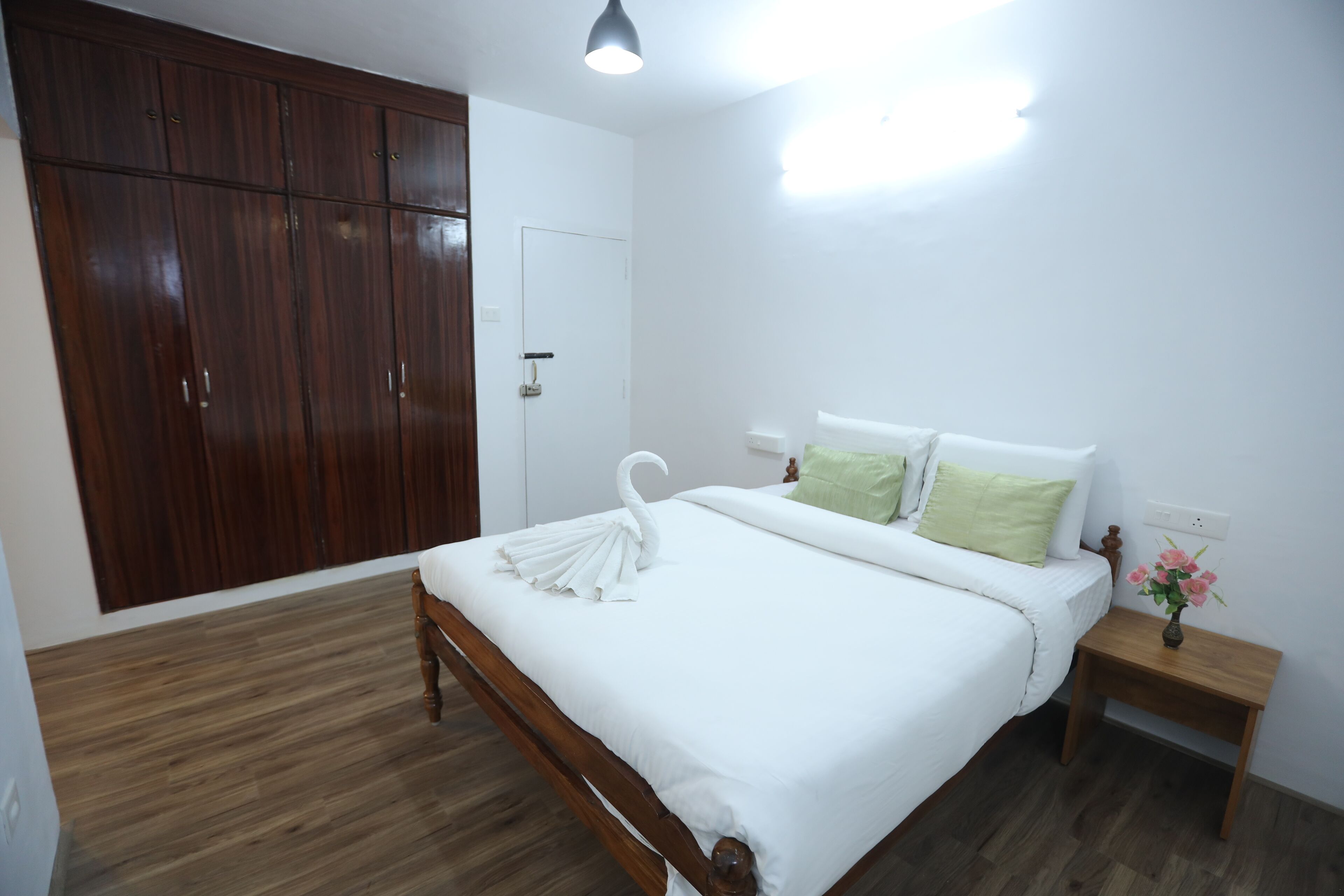 A bedroom with a wooden wardrobe, a bed with white bedding, a green pillow, and a nightstand with a vase of flowers.