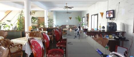 Une salle à manger spacieuse avec de hauts plafonds, des ventilateurs de plafond et une longue table entourée de chaises à revêtement rouge.