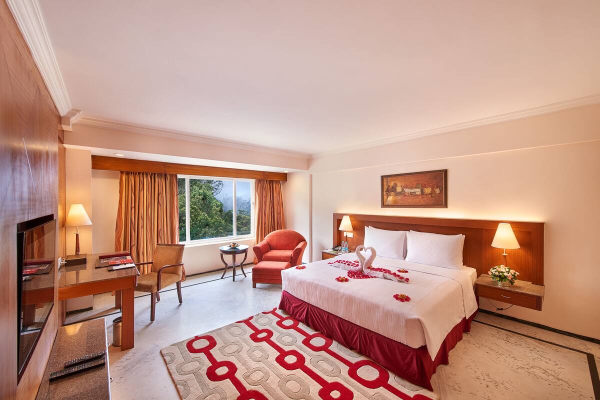 A hotel room with a large bed, a desk with a lamp, a chair, a small table, and a window with curtains.