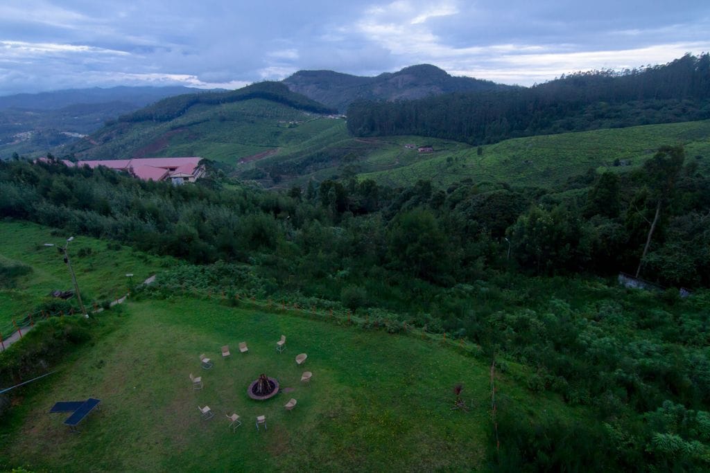 A grassy area with scattered chairs and a circular structure, surrounded by lush greenery and hills in the background.