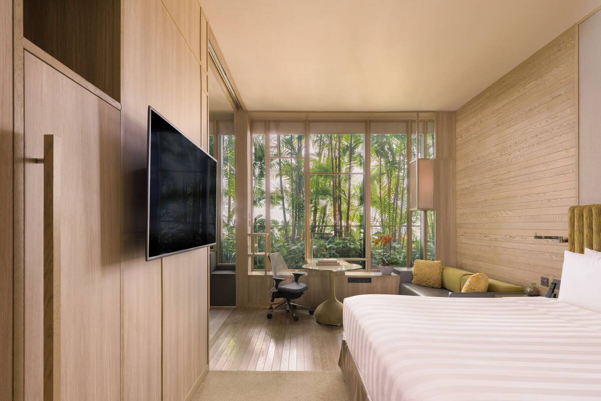 Une chambre d’hôtel moderne dotée d’un grand lit, d’un bureau avec une chaise, d’une télévision et de grandes fenêtres offrant une vue sur la végétation tropicale.