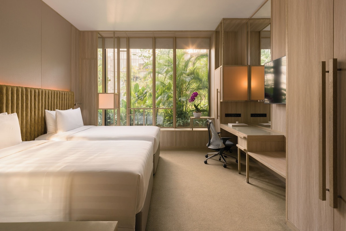 A hotel room with two beds, a desk, a chair, a TV, and a window with a view of greenery.