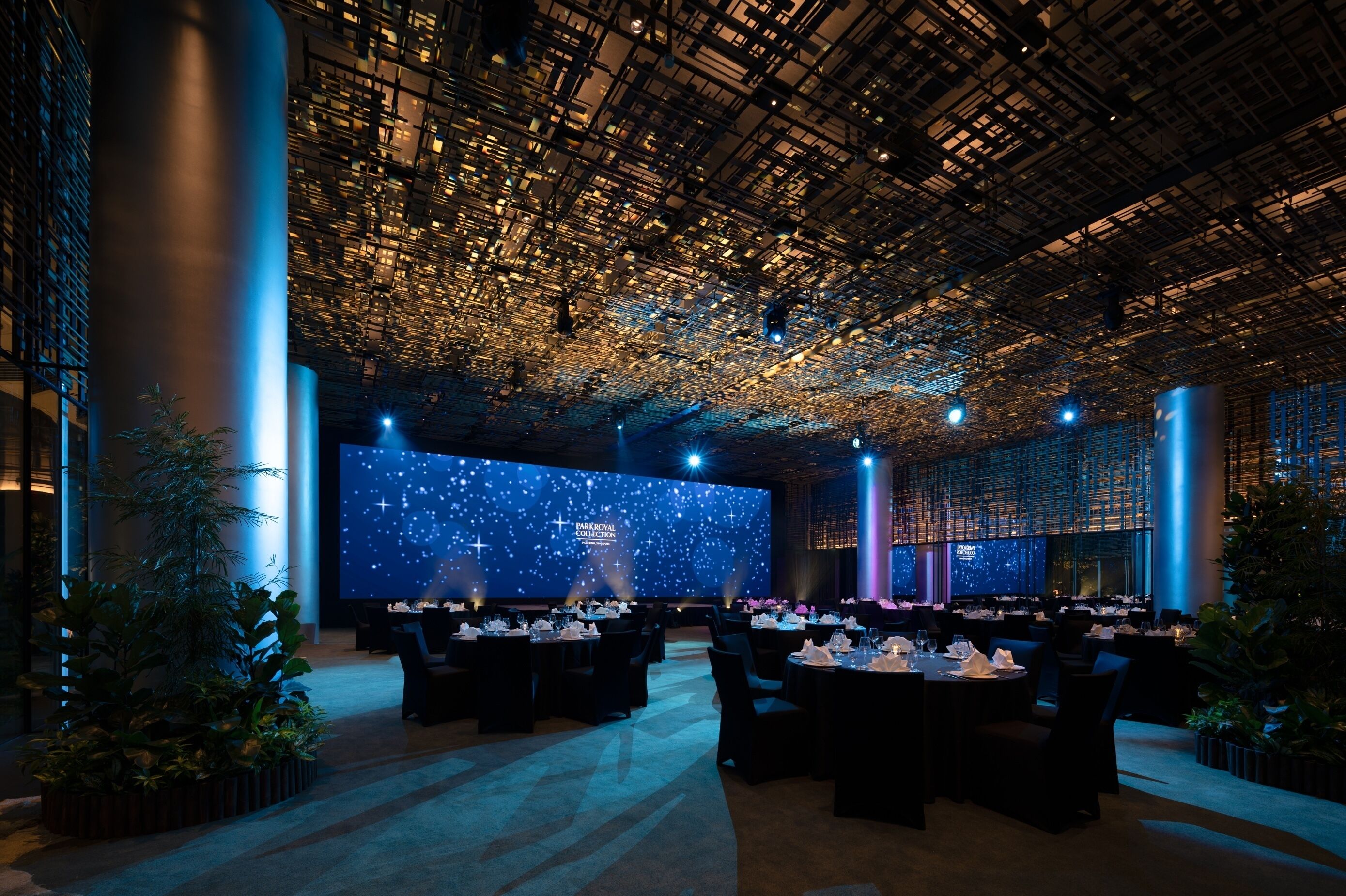 Un amplio salón de banquetes con techo alto, ventanas grandes y una proyección de estrellas en la pared del fondo. Mesas dispuestas para un evento formal.