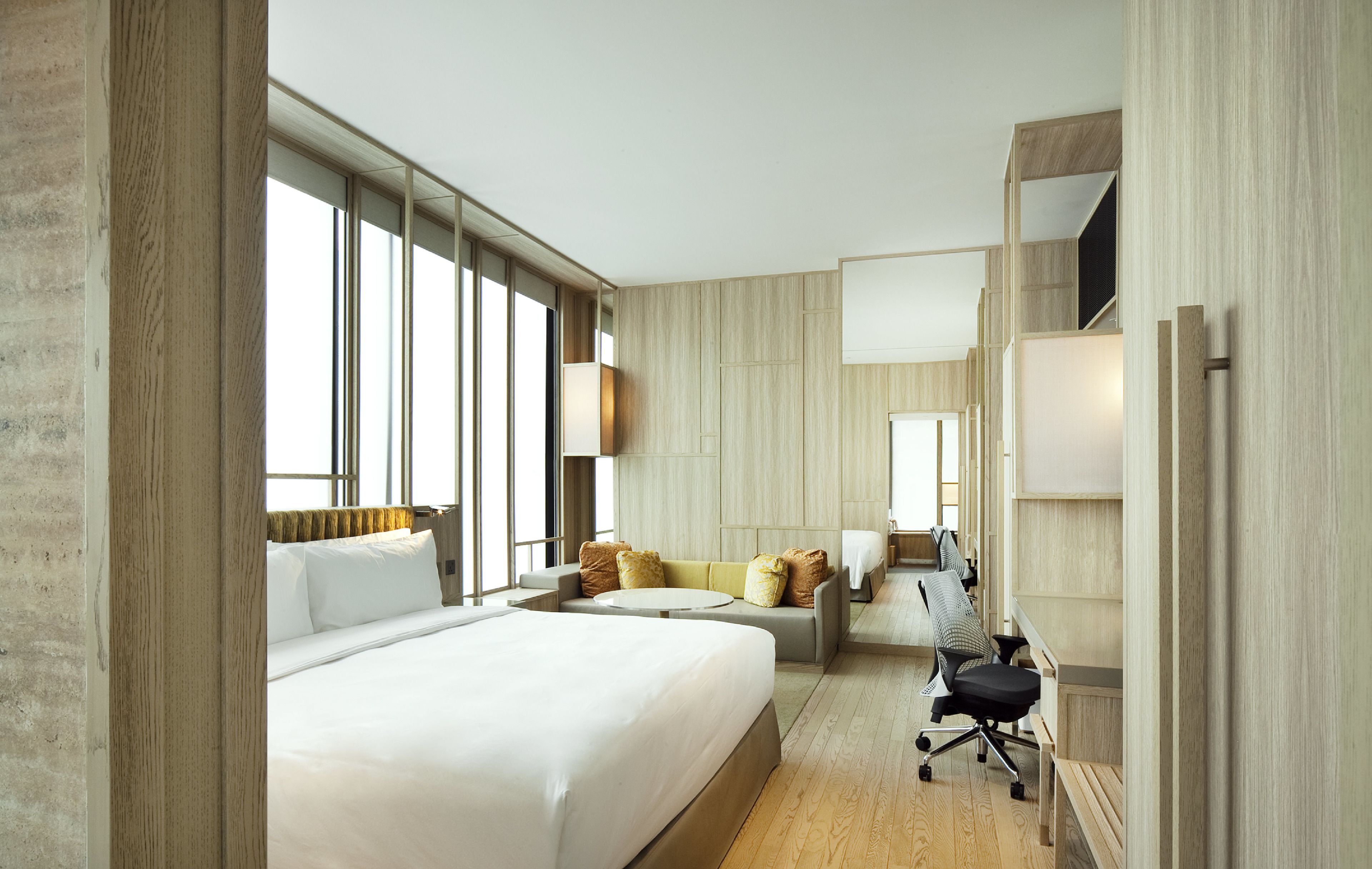 Une chambre d’hôtel moderne dotée d’un grand lit, d’un canapé, d’un bureau avec une chaise et d’une grande fenêtre laissant entrer la lumière naturelle.