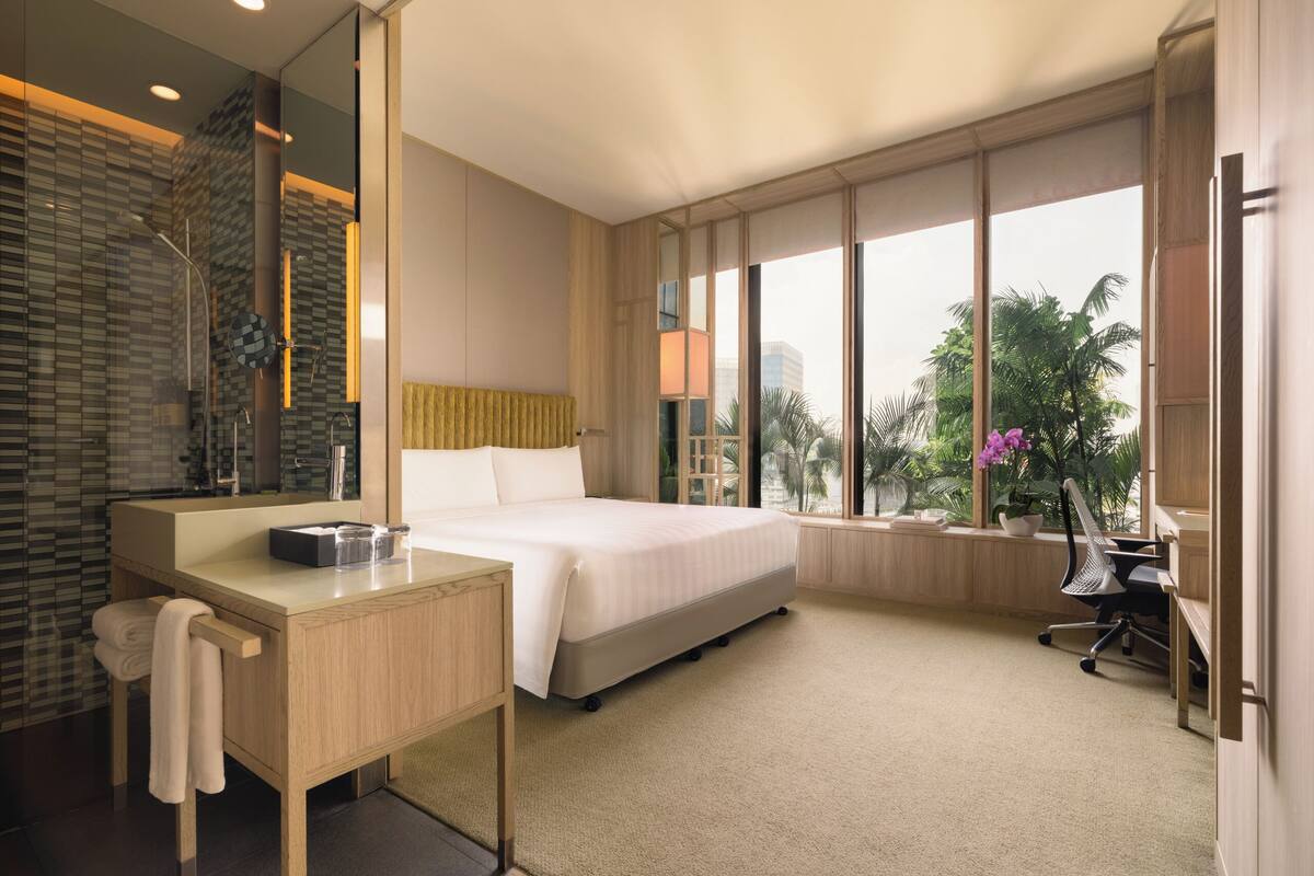 Une chambre d’hôtel moderne dotée d’un grand lit, d’un bureau avec un miroir, d’une salle de bain avec une douche en verre, et offrant une vue sur les palmiers à travers de grandes fenêtres.