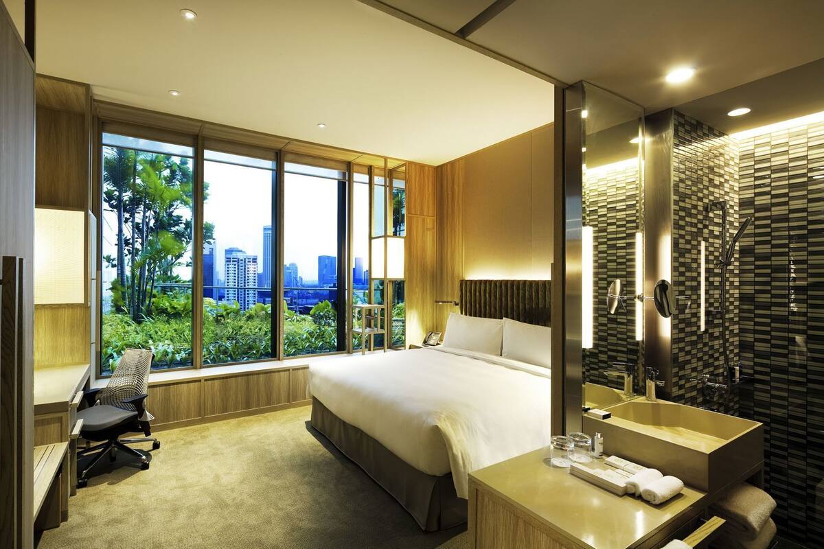 Une chambre d’hôtel moderne dotée d’un grand lit, d’un bureau, d’une chaise, d’une salle de bain avec un grand miroir et offrant une vue sur la ville grâce à de grandes fenêtres.