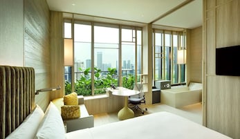 A modern hotel room with a large bed, a desk, and a view of a cityscape through large windows.