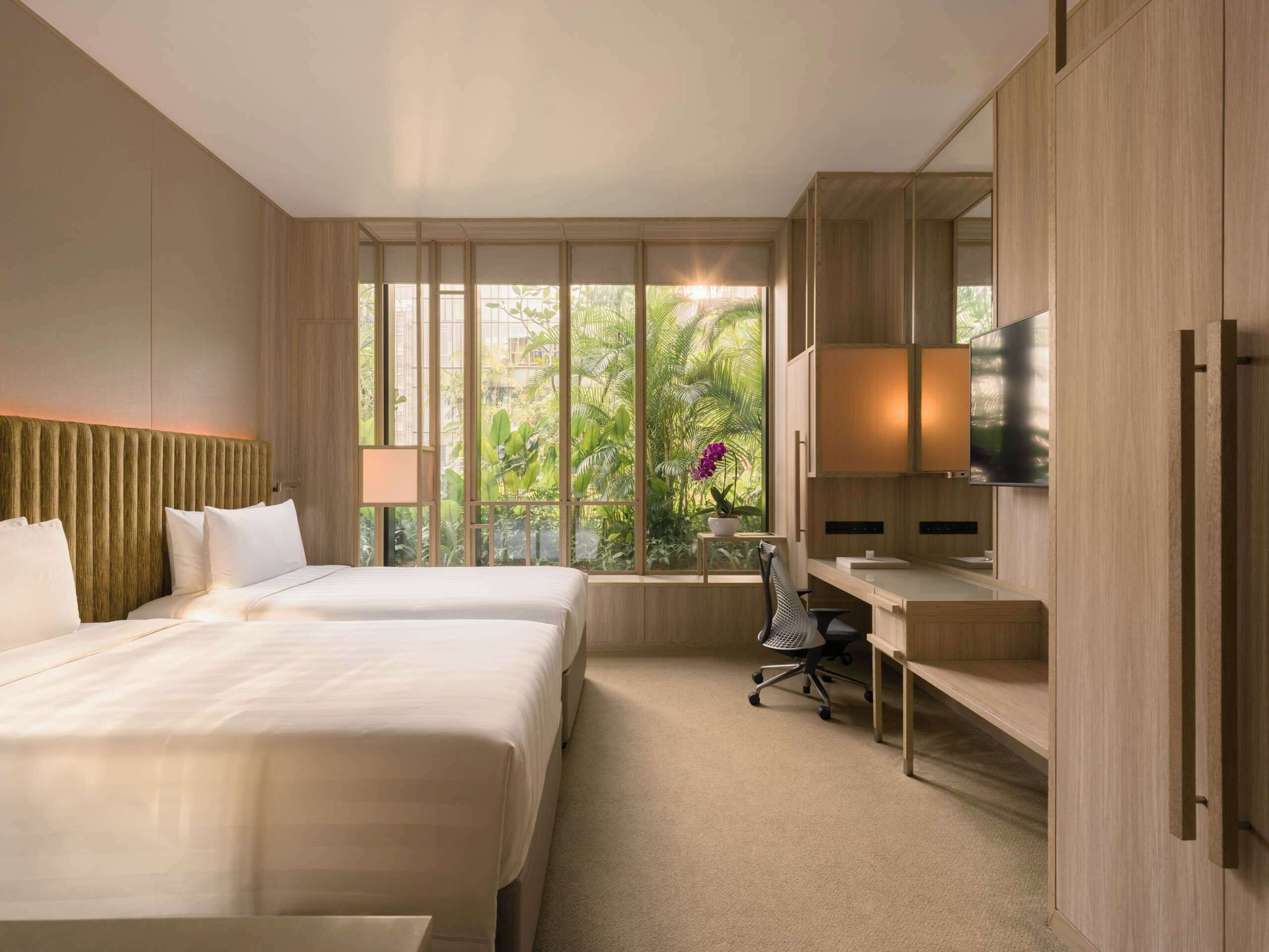 A hotel room with two beds, a desk, a chair, a TV, and a window with a view of greenery.