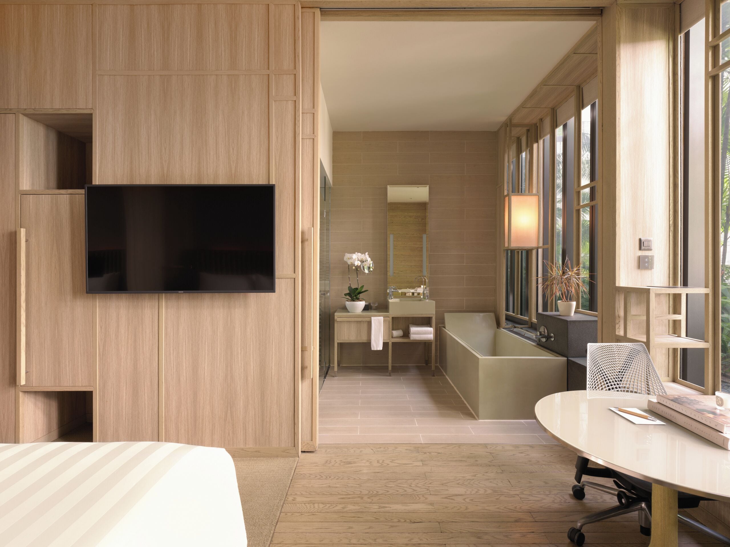 A modern hotel room with a large flat-screen TV mounted on the wall, a wooden wardrobe, a bathroom with a bathtub, a round table with a chair, and large windows providing natural light.