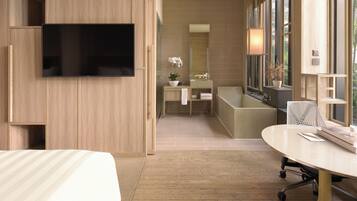A modern hotel room with a large flat-screen TV mounted on the wall, a wooden wardrobe, a bathroom with a bathtub, a round table with a chair, and large windows providing natural light.