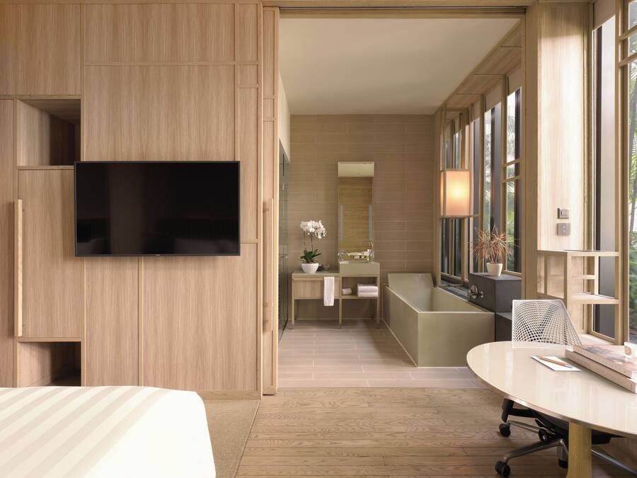A modern hotel room with a large flat-screen TV mounted on the wall, a wooden wardrobe, a bathroom with a bathtub, a round table with a chair, and large windows providing natural light.
