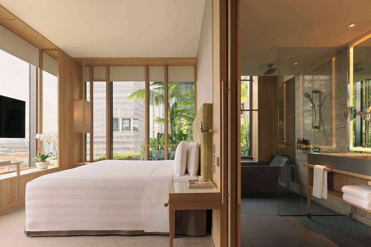 Une chambre d’hôtel moderne dotée d’un grand lit, d’une table de chevet, d’un miroir, d’une salle de bain avec douche et de grandes fenêtres offrant une vue sur les palmiers et les immeubles.