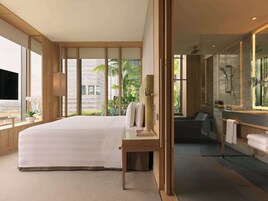 A modern hotel room with a large bed, a bedside table, a mirror, a bathroom with a shower, and large windows offering a view of palm trees and buildings.