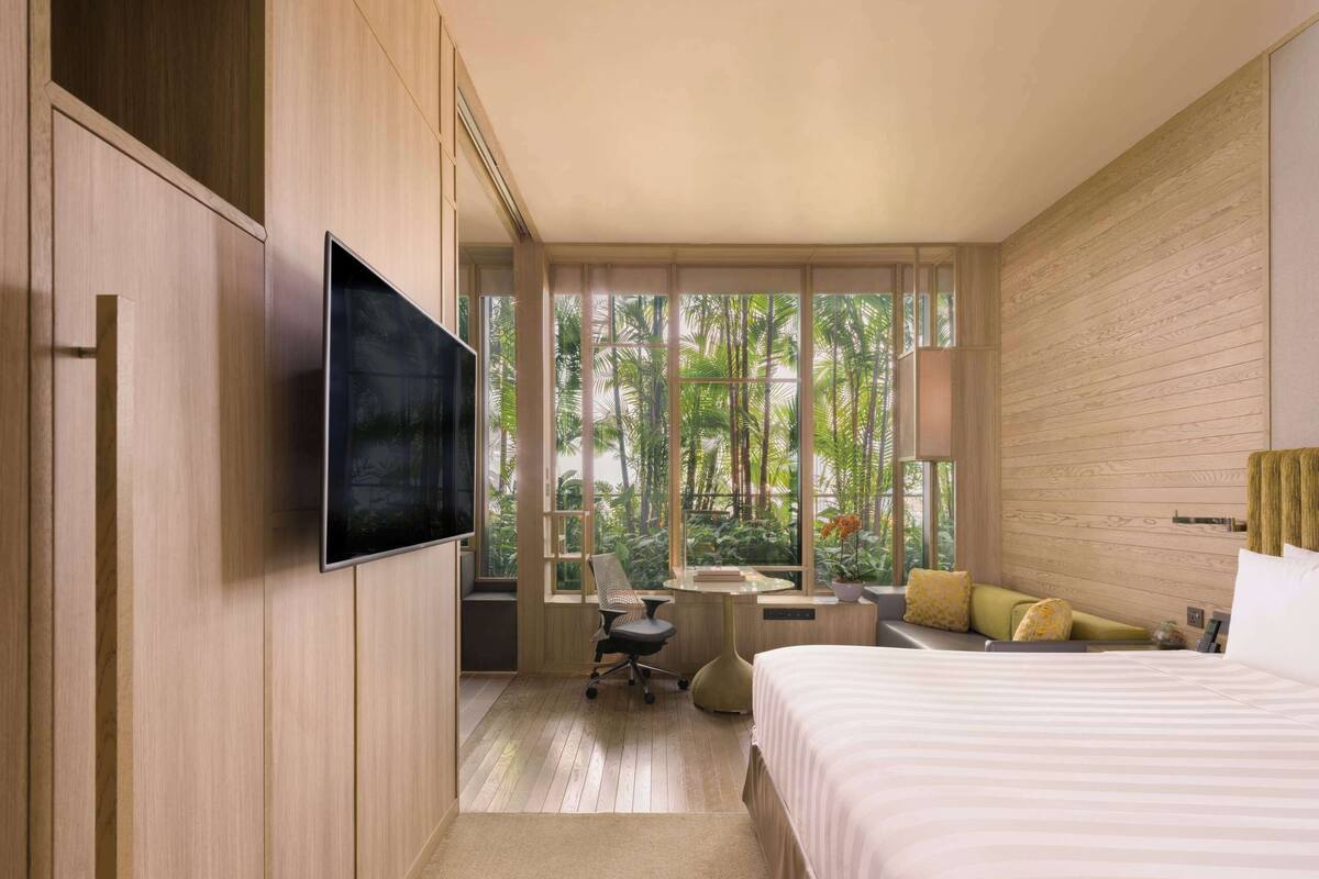 Une chambre d’hôtel moderne dotée d’un grand lit, d’un bureau avec une chaise, d’une télévision et de grandes fenêtres offrant une vue sur la végétation tropicale.