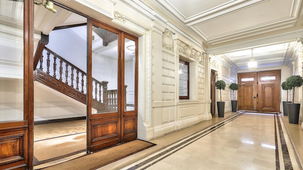 Un amplio pasillo con escaleras de madera, puertas grandes y piso de mármol.