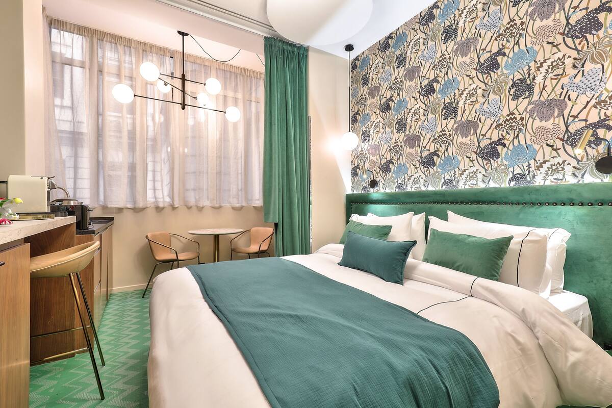 A hotel room with a bed, a desk, a chair, a small table, and a patterned wall.