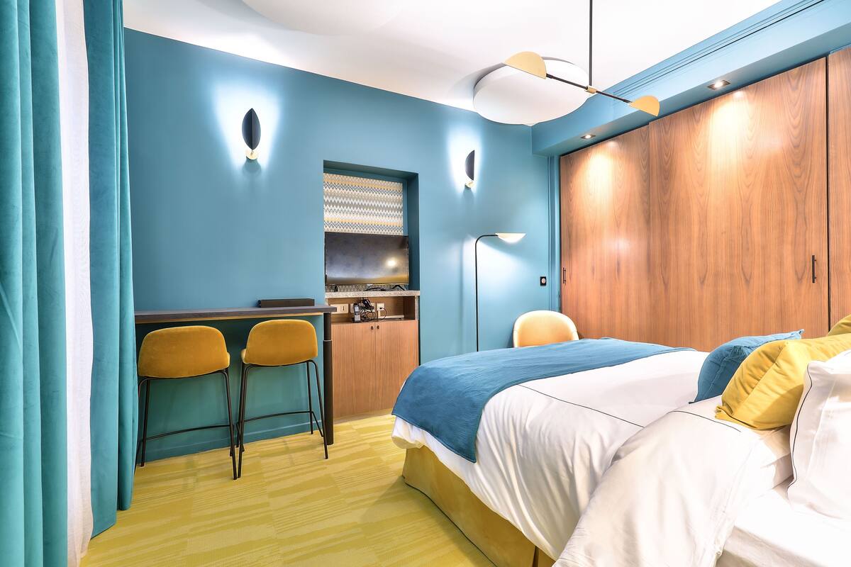 A hotel room with a bed, a desk with two chairs, a wardrobe, and a small kitchenette.