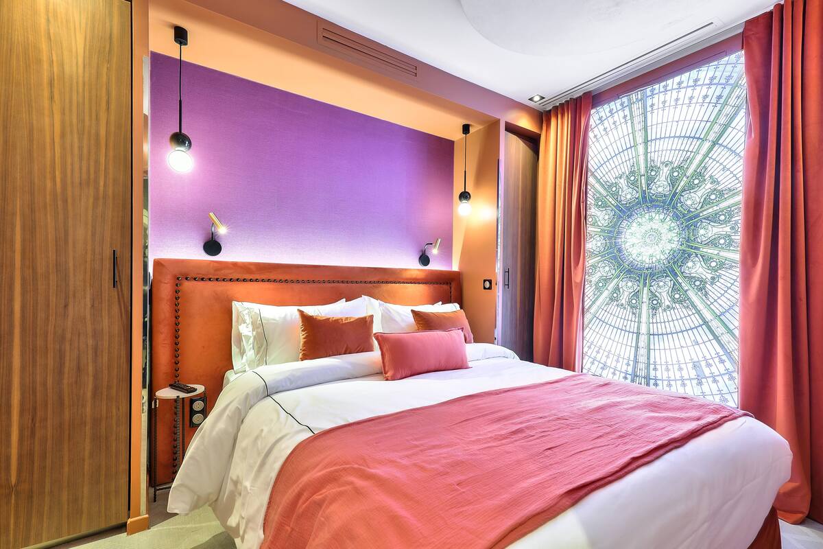 A hotel room with a large bed, a wooden wardrobe, a small bedside table, and a decorative window with curtains.