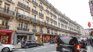 Vista de una calle con una hilera de edificios de varios pisos, comercios y coches aparcados.