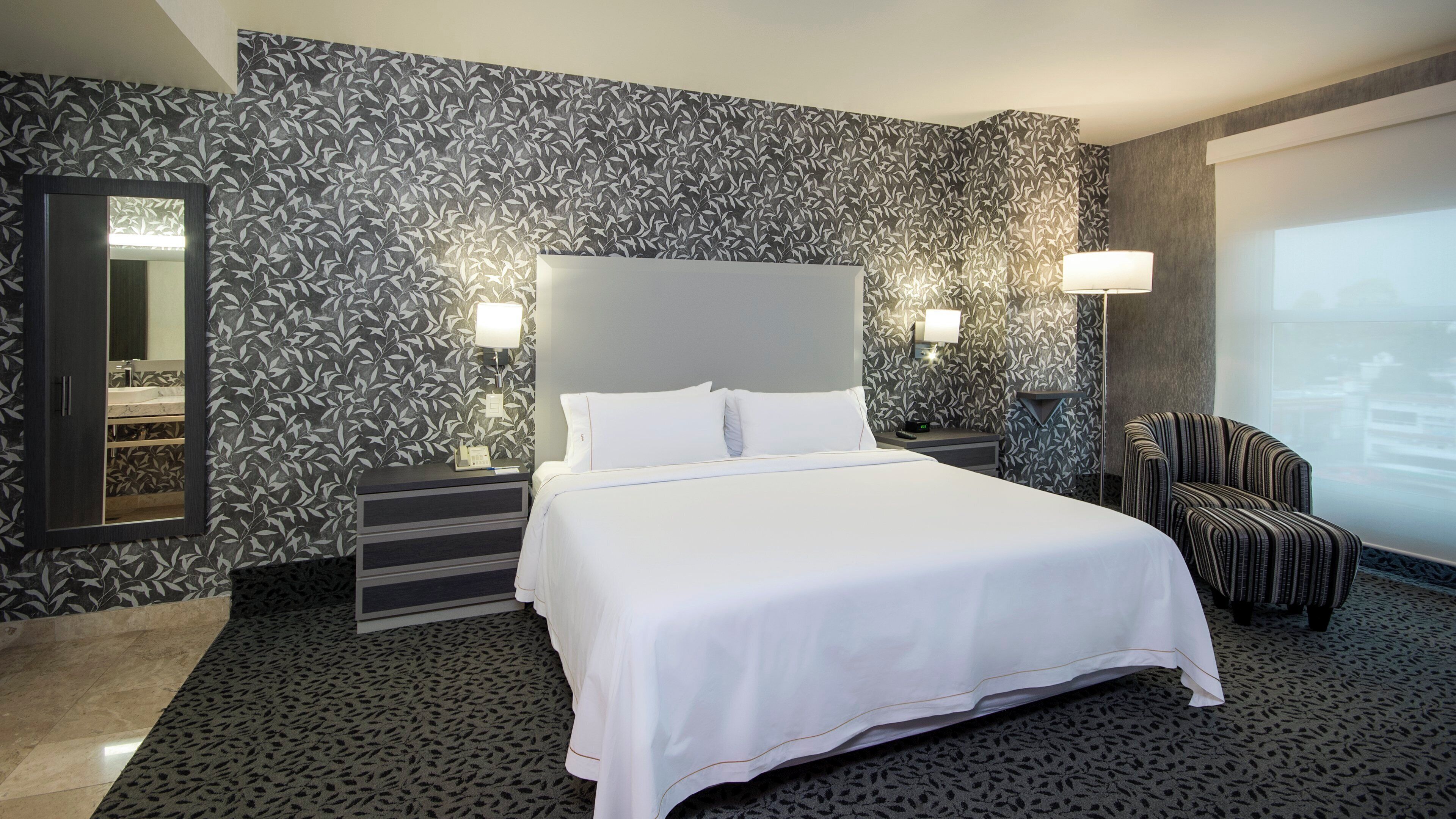 Une chambre d’hôtel avec un grand lit, une table de chevet, une chaise, un miroir et un papier peint à motifs.
