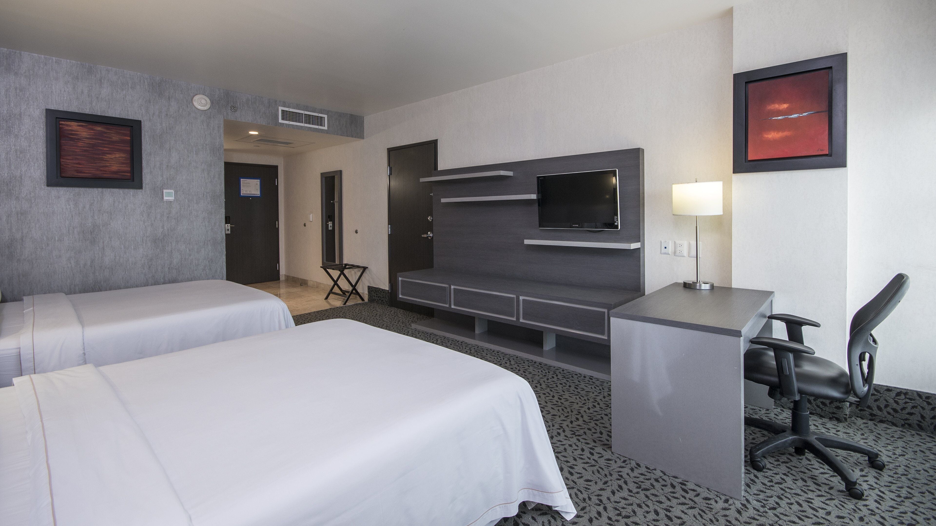 Quarto de hotel com duas camas, uma escrivaninha com cadeira, televisão de tela plana, abajur e obras de arte nas paredes.