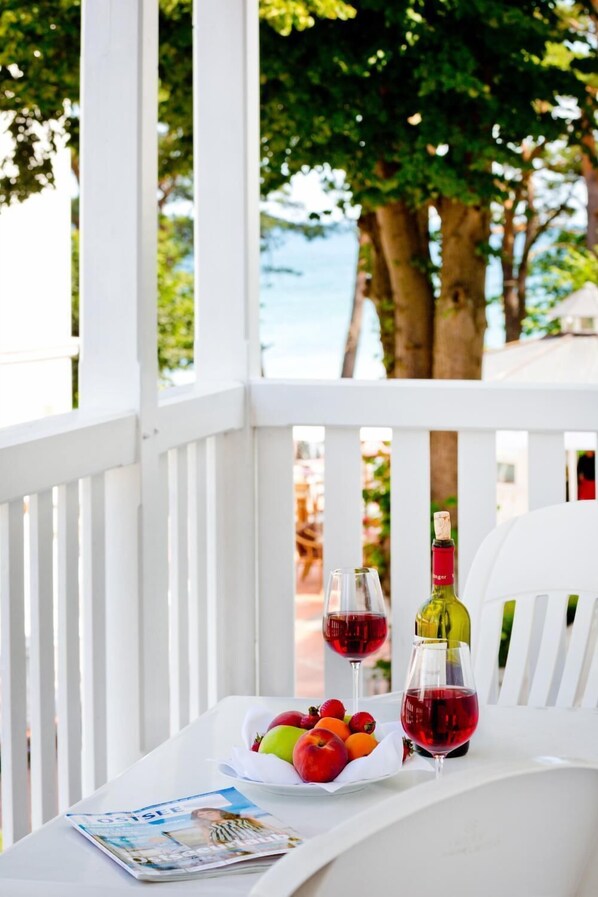 Una mesa con una botella de vino, copas y un frutero, sobre una mesa blanca en un espacio al aire libre cubierto, con árboles y vistas al mar.