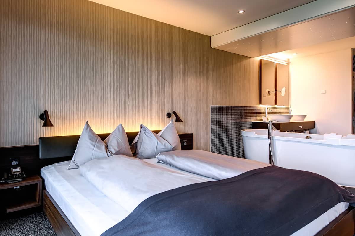 A modern hotel room with a large bed, a built-in bathtub, and a mirror above the sink.