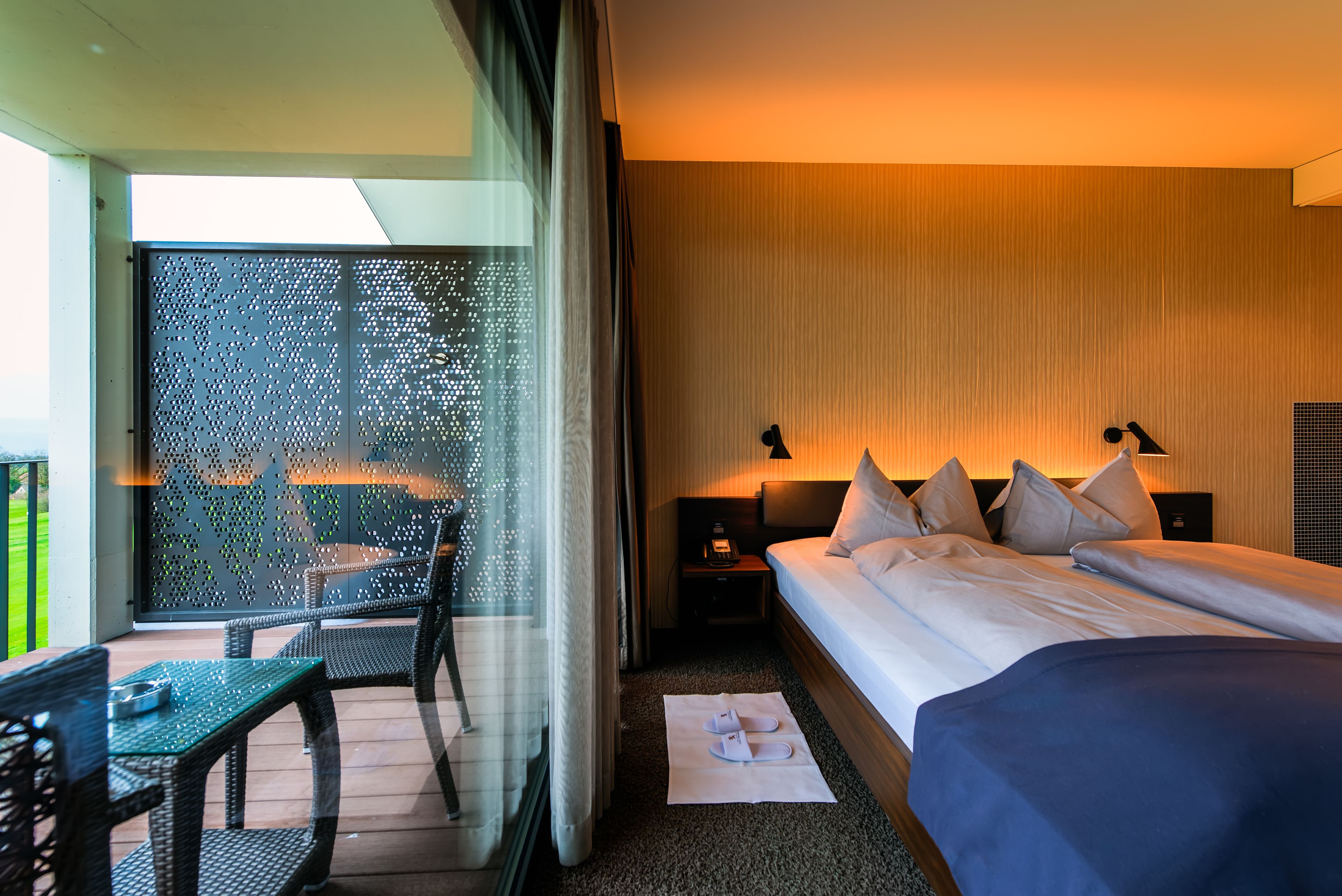 A hotel room with a balcony, a bed with multiple pillows, a nightstand with a lamp, and a view through a patterned glass door.