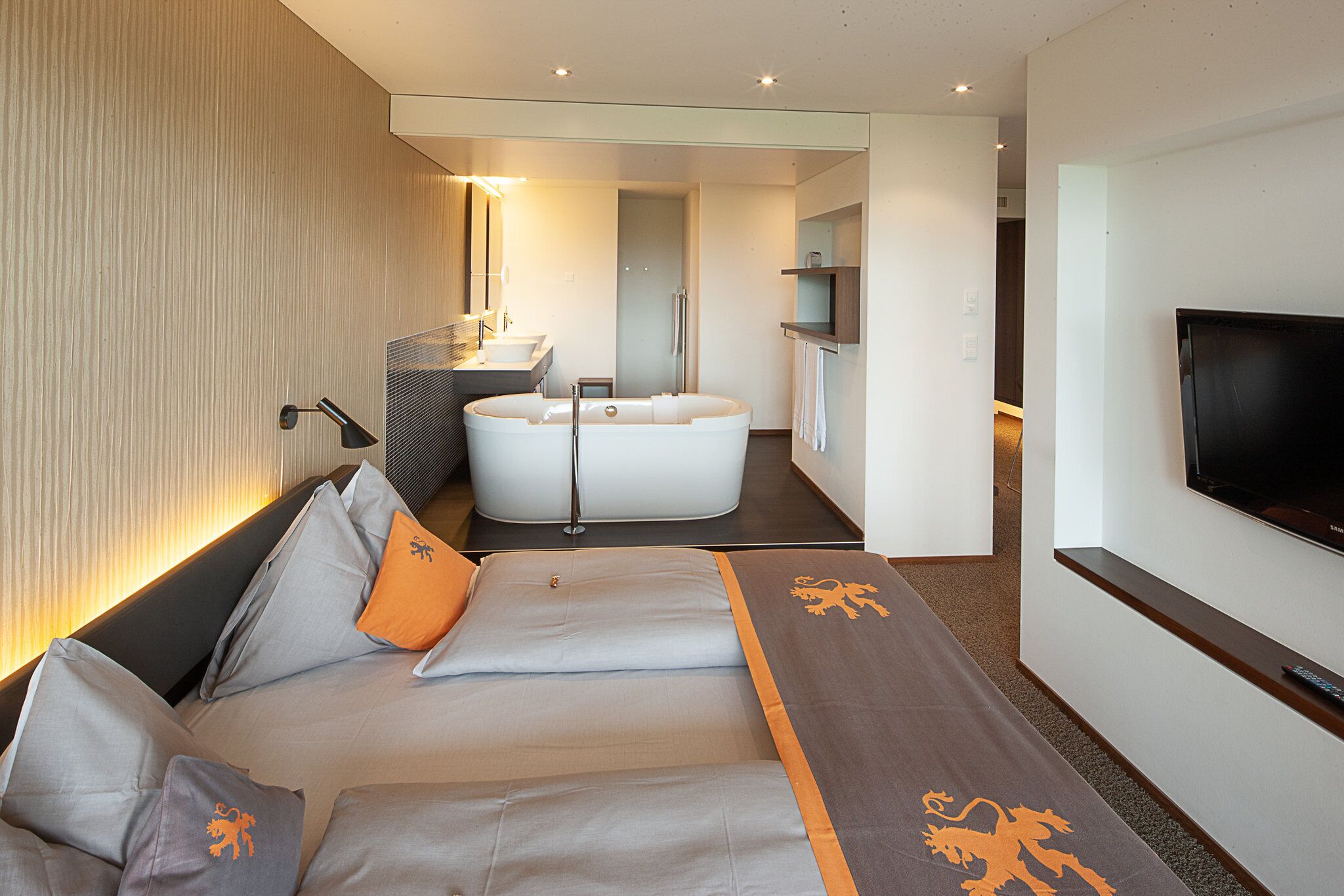 A modern hotel room with a sofa, a bathtub, a TV, and a bathroom with a mirror and sink.