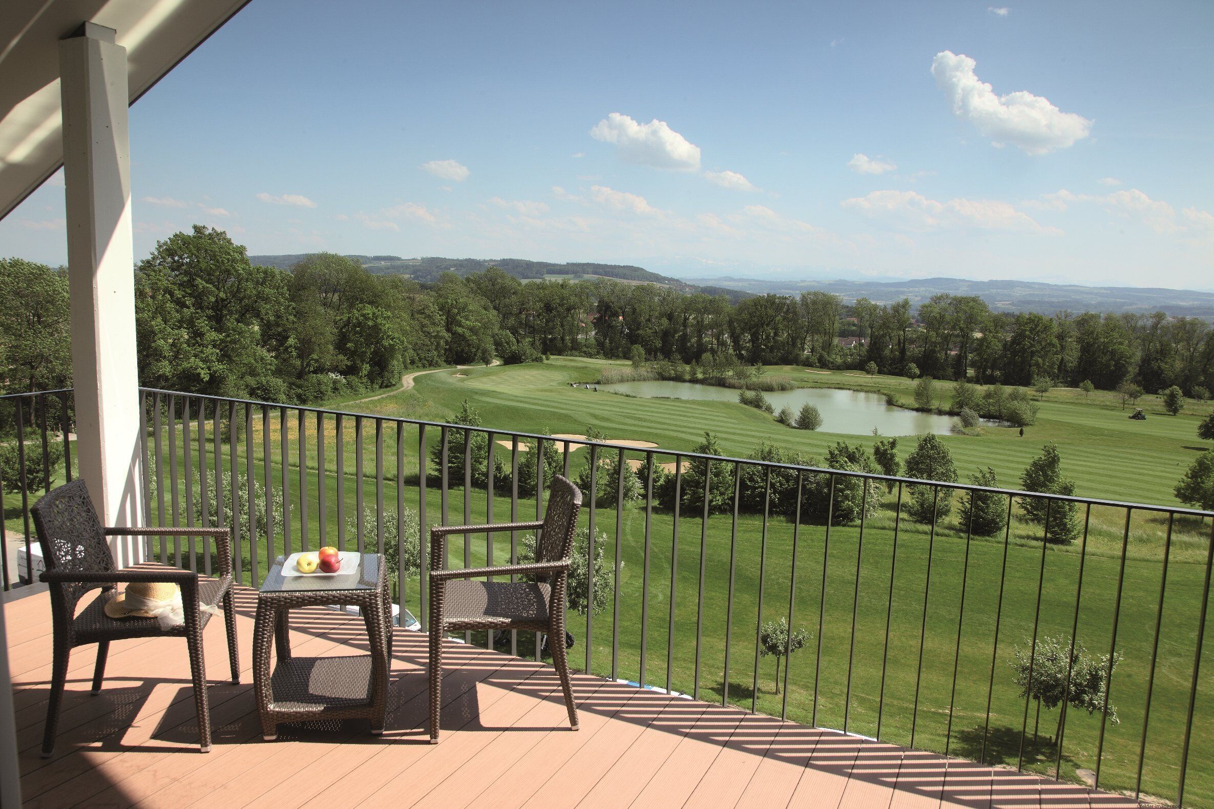 A balcony with wicker chairs and a table, overlooking a golf course and a lake.