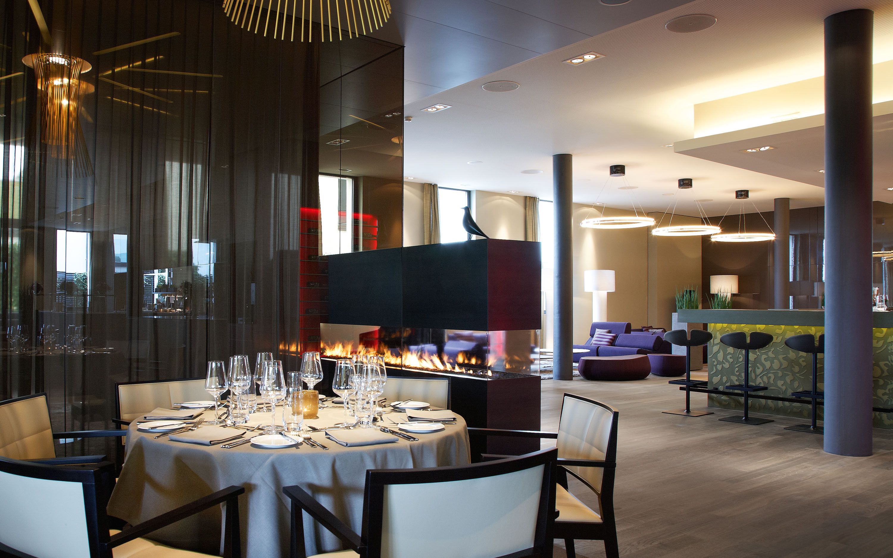 Ein Restaurant mit einem zentralen Kamin, runden Tischen und moderner Einrichtung.