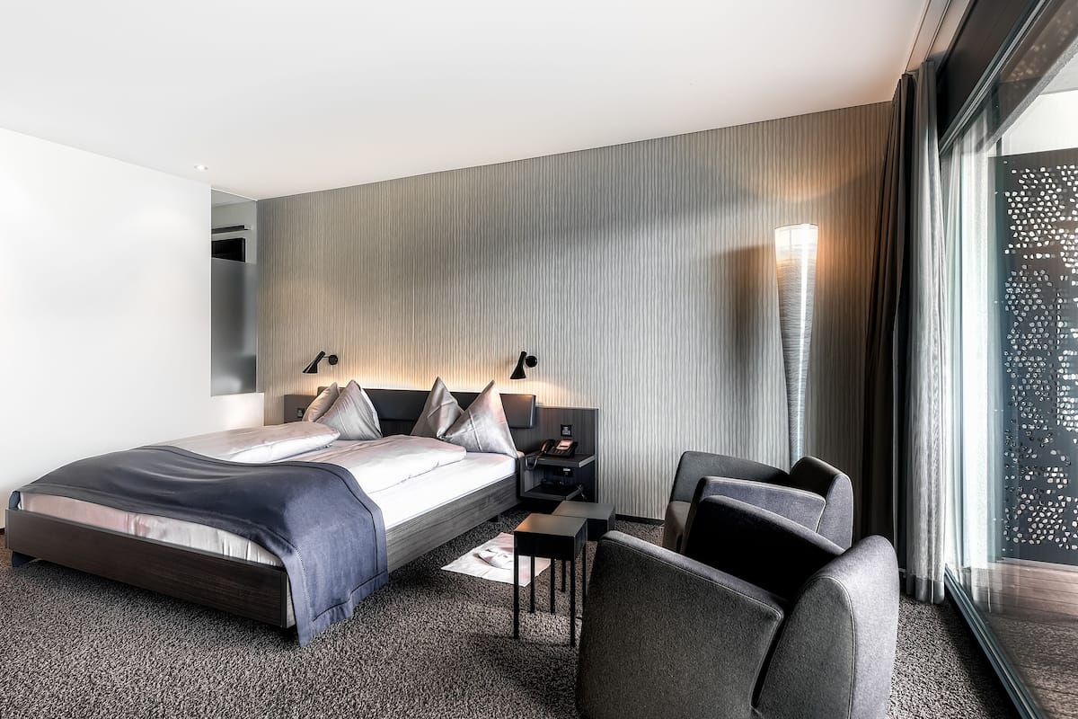 A modern hotel room with a bed, bedside table, chair, and a large window with a view.