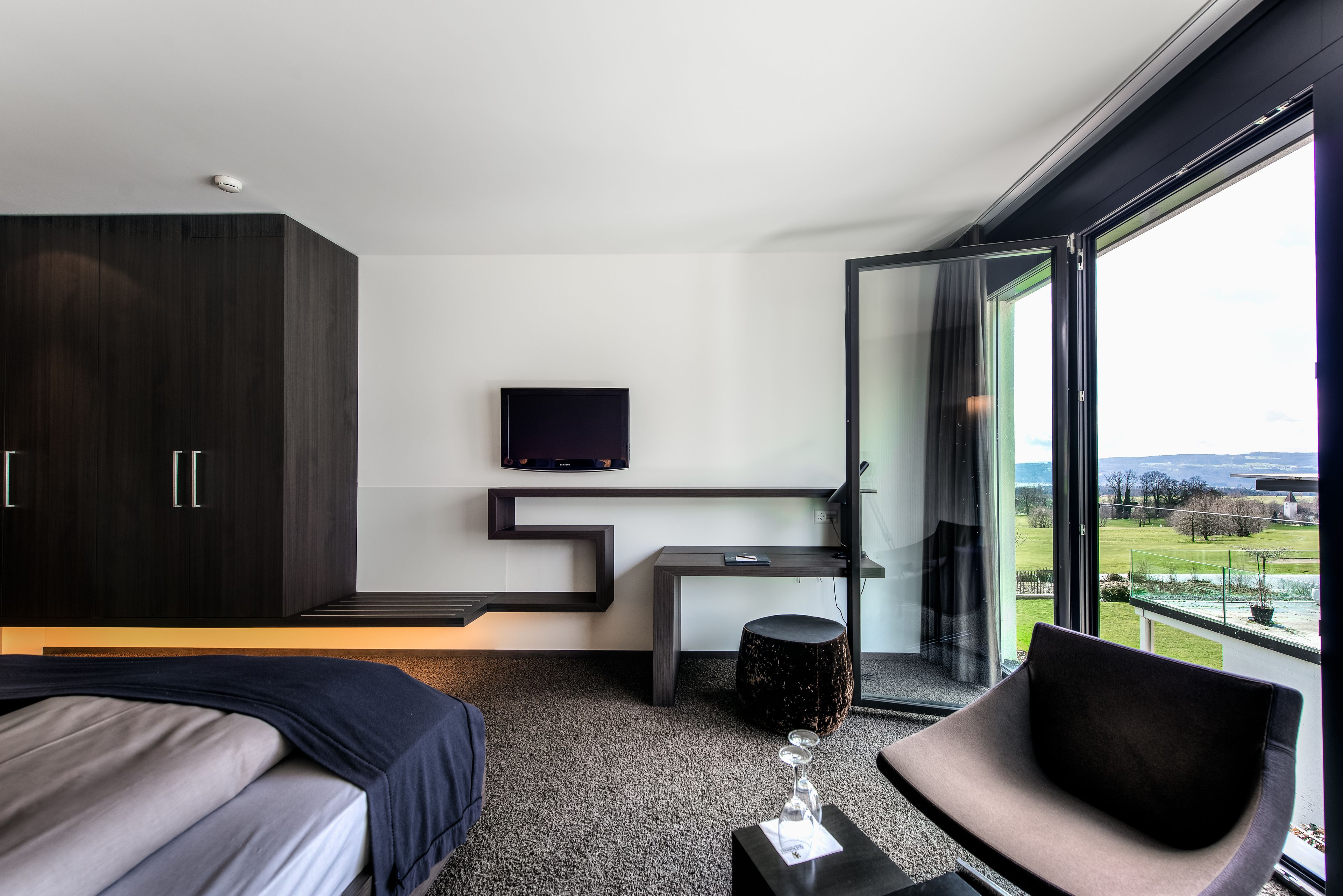 Une chambre d’hôtel moderne équipée d’un lit, d’un canapé, d’un téléviseur fixé au mur et d’une grande fenêtre donnant sur l’extérieur.