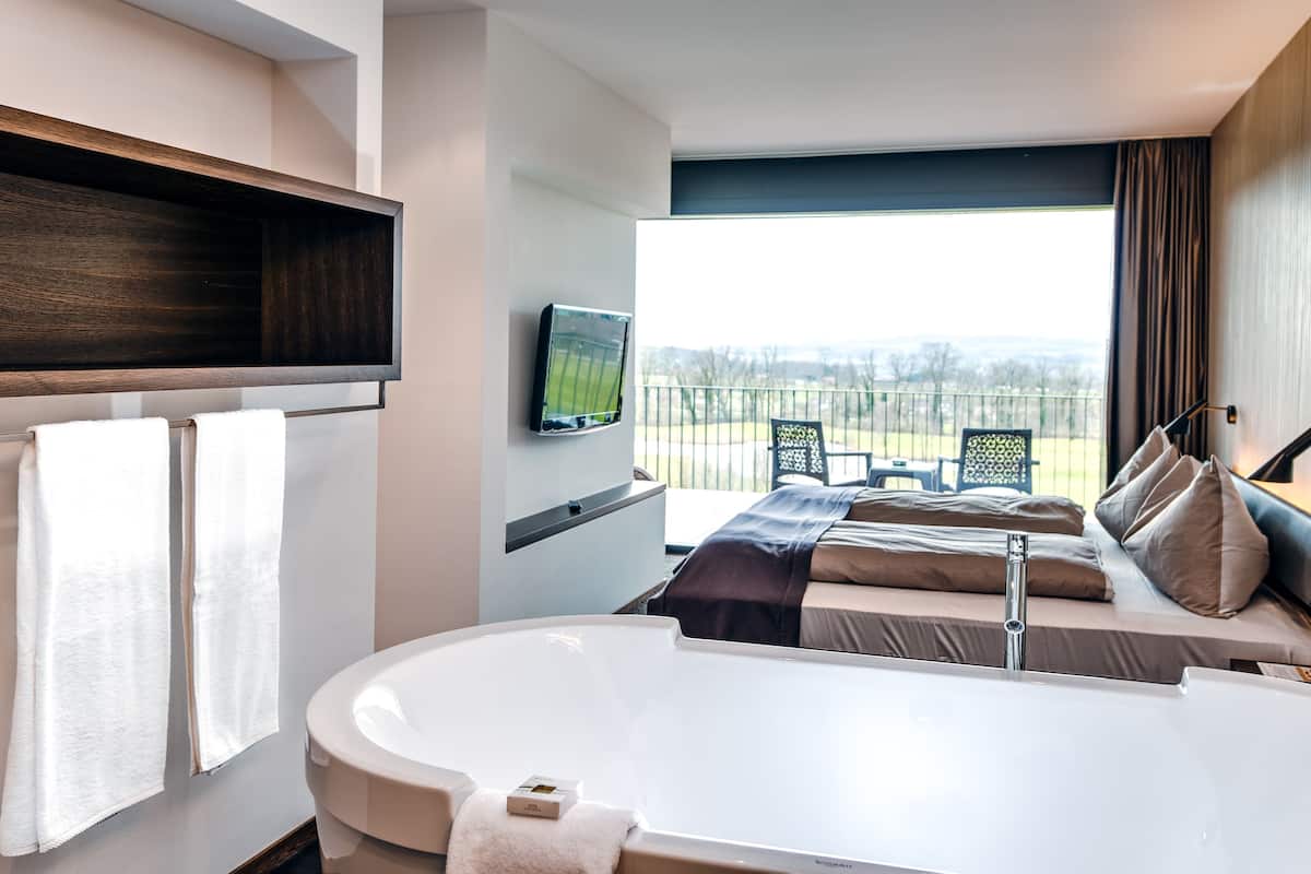 A modern hotel room with a large bathtub, a bed with multiple pillows, a television mounted on the wall, and a balcony with outdoor furniture.