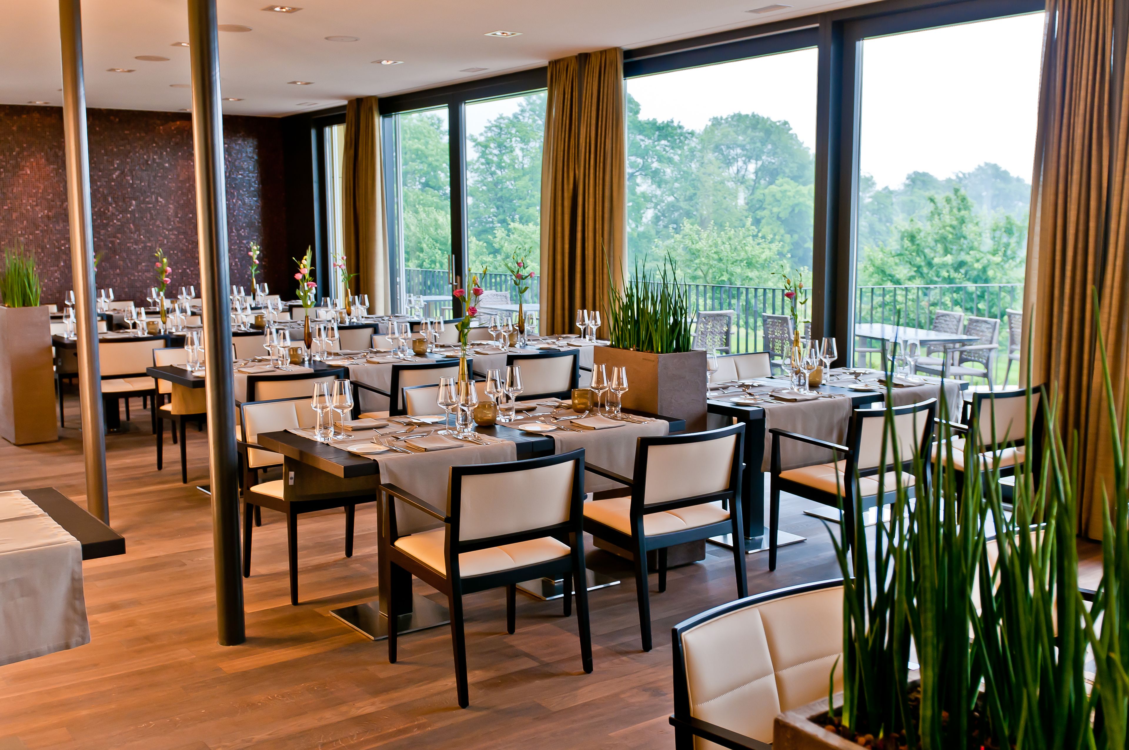 En restaurant med flere spiseborde, store vinduer med udsigt til grønne omgivelser og moderne indretning.