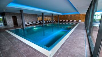 Ein moderner, überdachter Swimmingpool mit klarem, blauem Wasser, gefliestem Boden und einer Reihe von Liegestühlen am Rand.