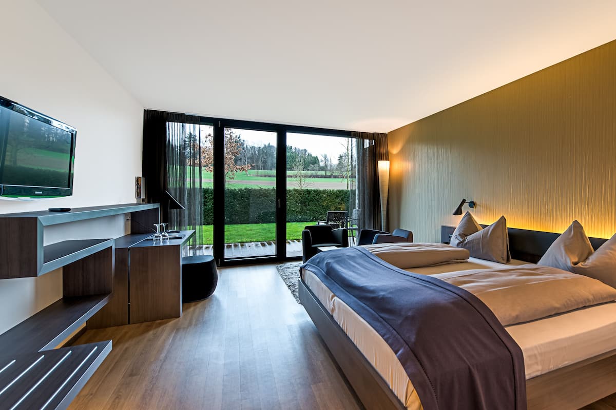 A modern hotel room with a large bed, a desk with a flat-screen TV, and a sliding glass door leading to a garden.