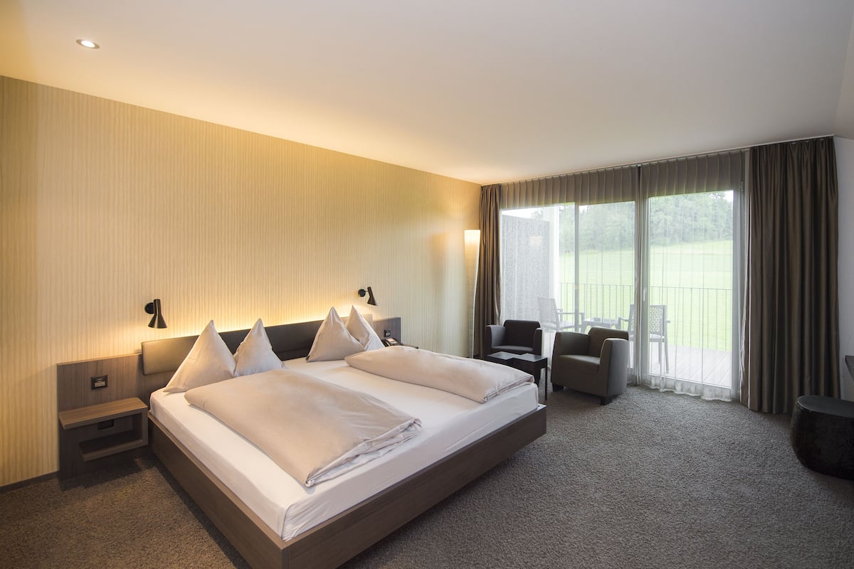 A hotel room with a large bed, a desk, a chair, and a view of a grassy area through the window.