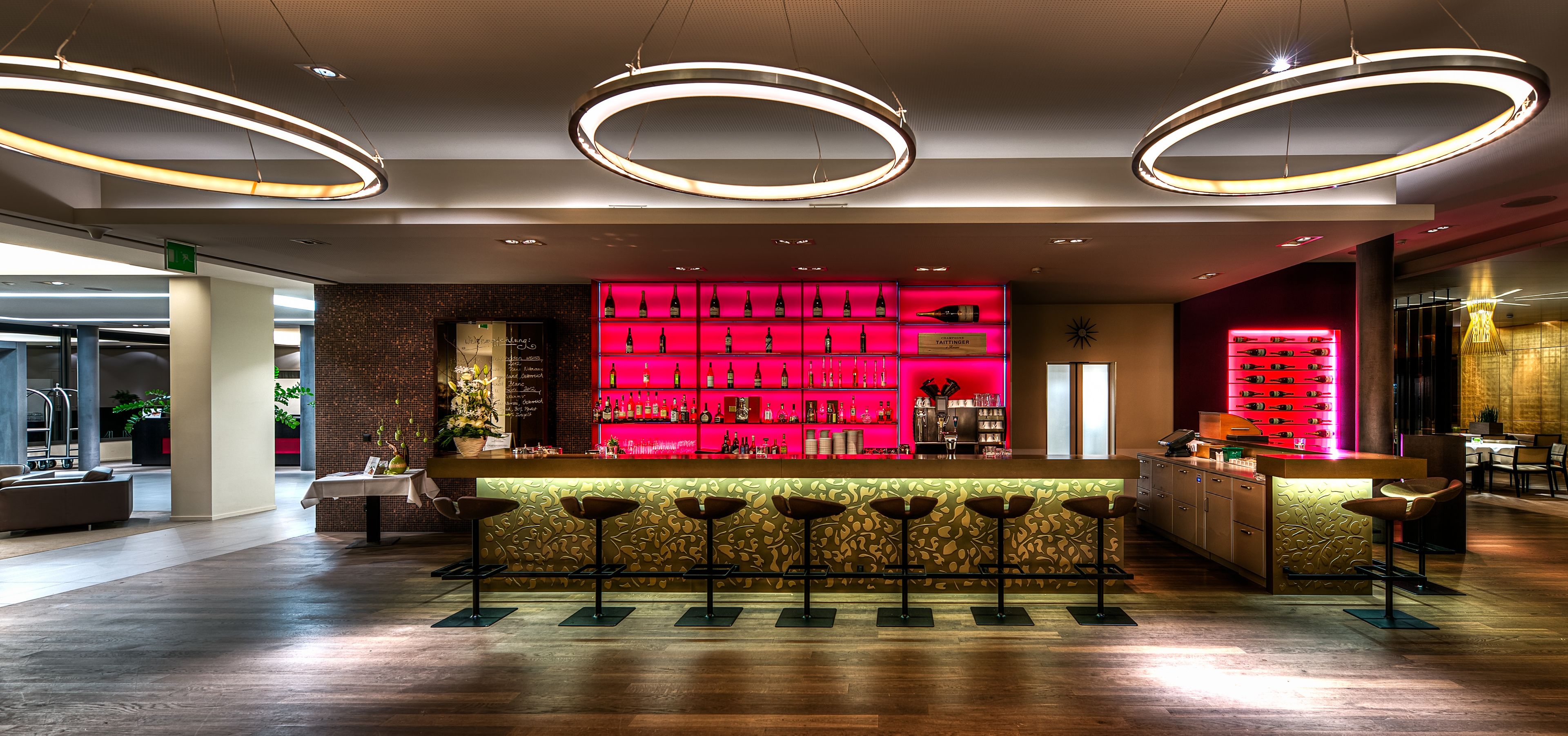 Een moderne bar met een roze plank verlicht door neon, waarop flessen staan, een houten toog en meerdere barstoelen.