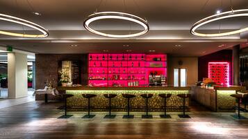 Een moderne bar met een roze plank verlicht door neon, waarop flessen staan, een houten toog en meerdere barstoelen.