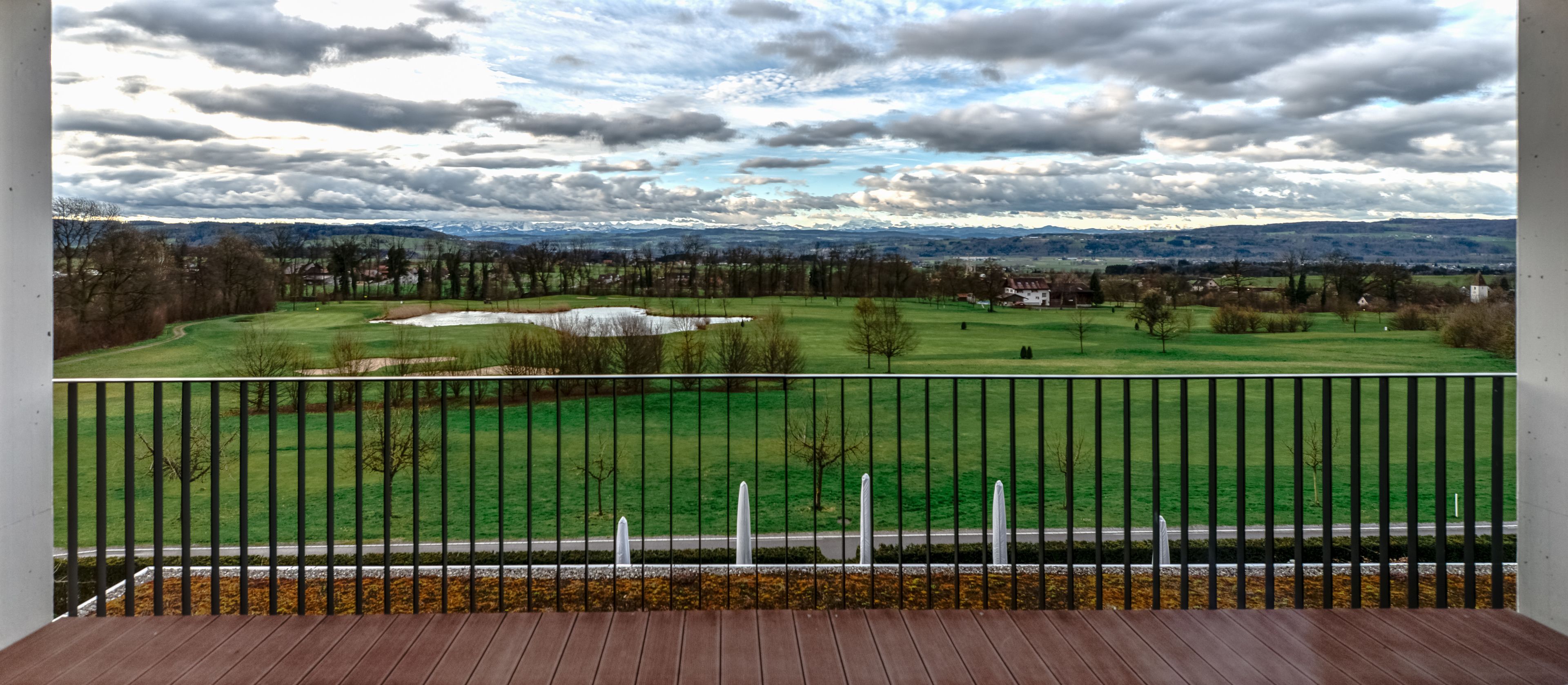 A balcony with a view of a golf course, a pond, and distant mountains.