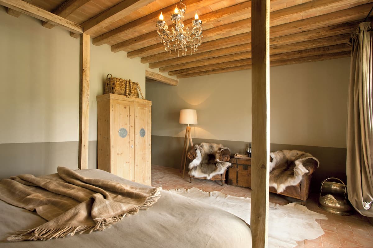 Una habitación con cama, vigas de madera en el techo, candelabro, aparador, sillón de piel y lámpara.