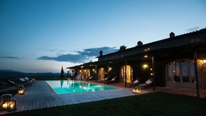 Un área junto a la piscina con terraza de madera, sillas de descanso y un edificio de piedra con ventanas de arco.