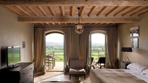 Un dormitorio con una cama grande, una silla, un escritorio y un televisor. Hay dos ventanas con cortinas, una araña y vistas al campo.