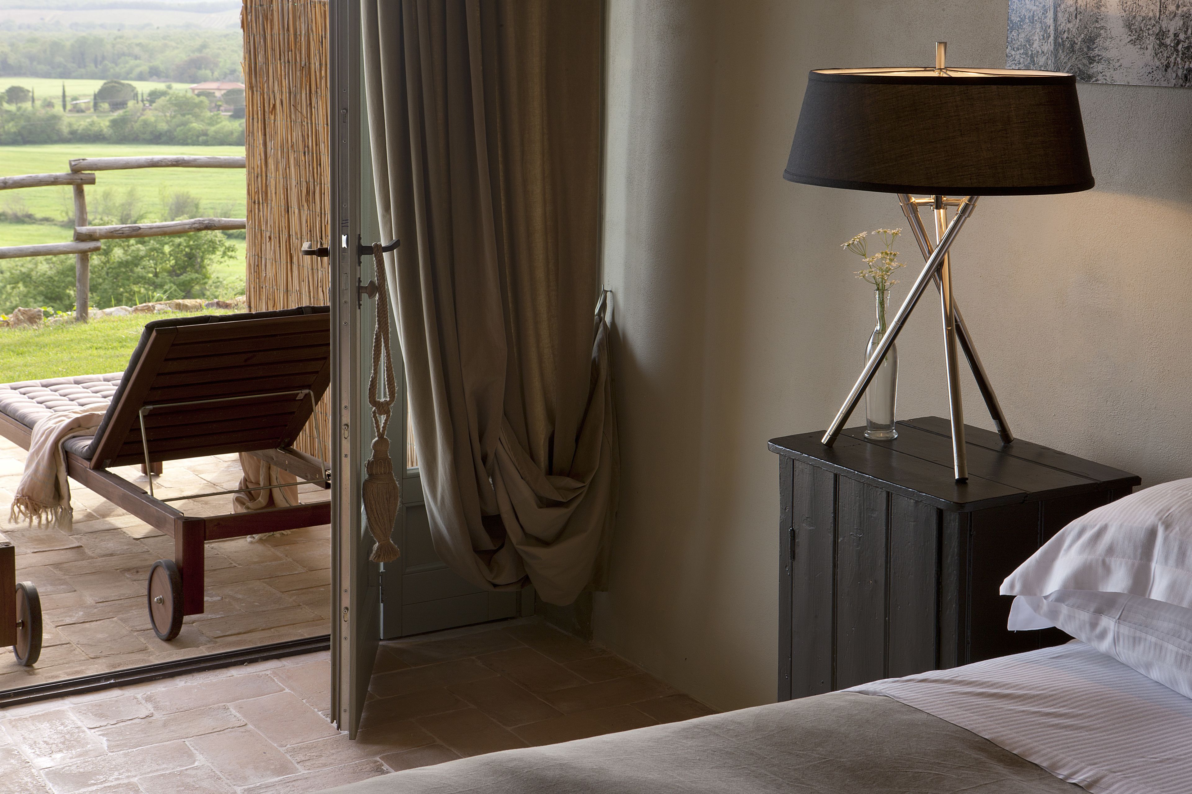 Un dormitorio con cama, mesita de noche, lámpara, silla y vistas al exterior.