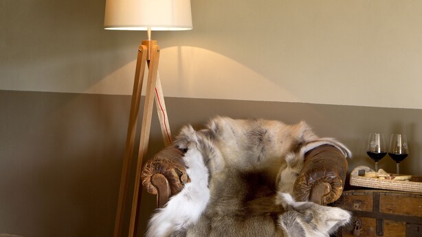 A room with a wooden floor, a lamp, a fur chair, and a wooden cabinet with wine glasses. - Conti di San Bonifacio Wine Resort (Gavorrano)