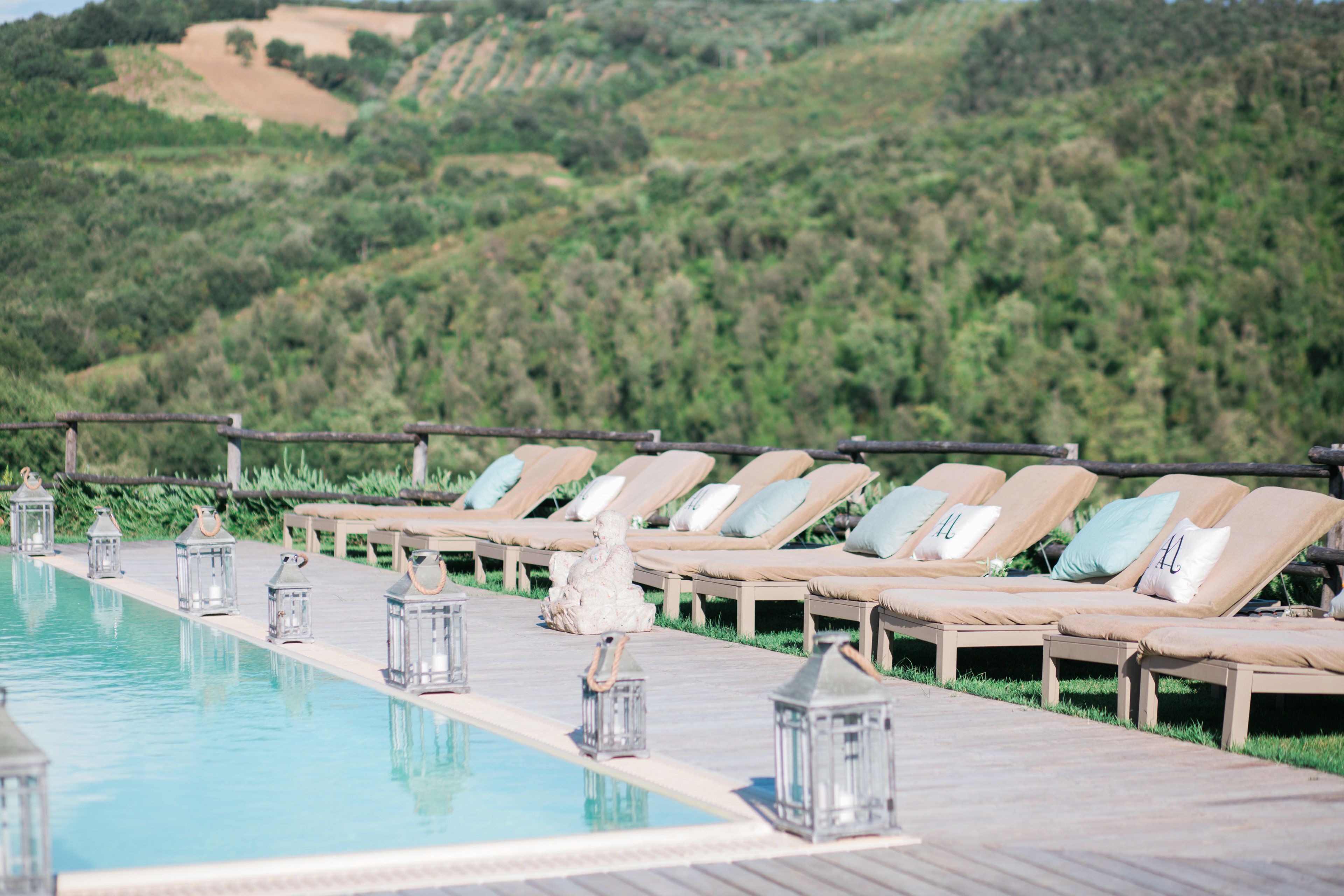 Zona junto a la piscina con sillas de descanso y faroles, con colinas verdes al fondo.