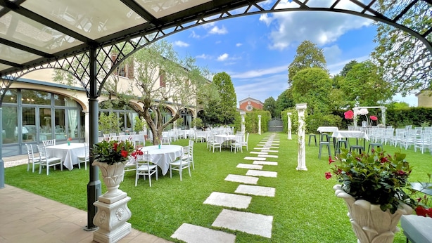 An outdoor garden with white tables and chairs arranged in rows, a stone pathway, and a pergola structure. - HC Hotel Il Gelso Pontevico (Pontevico)