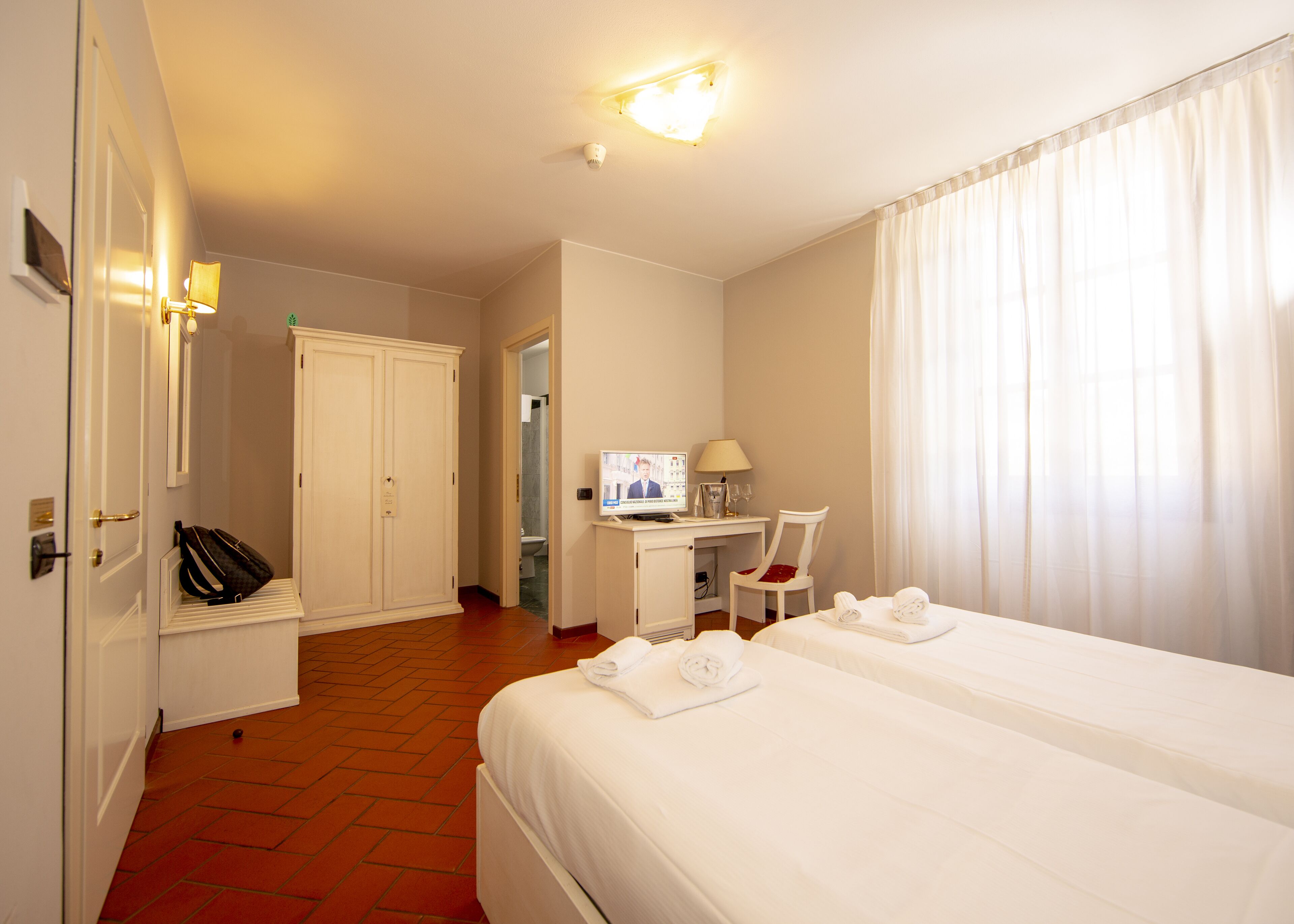 Quarto de hotel com duas camas, uma escrivaninha com computador, uma cadeira, televisão e banheiro visível por uma porta aberta.