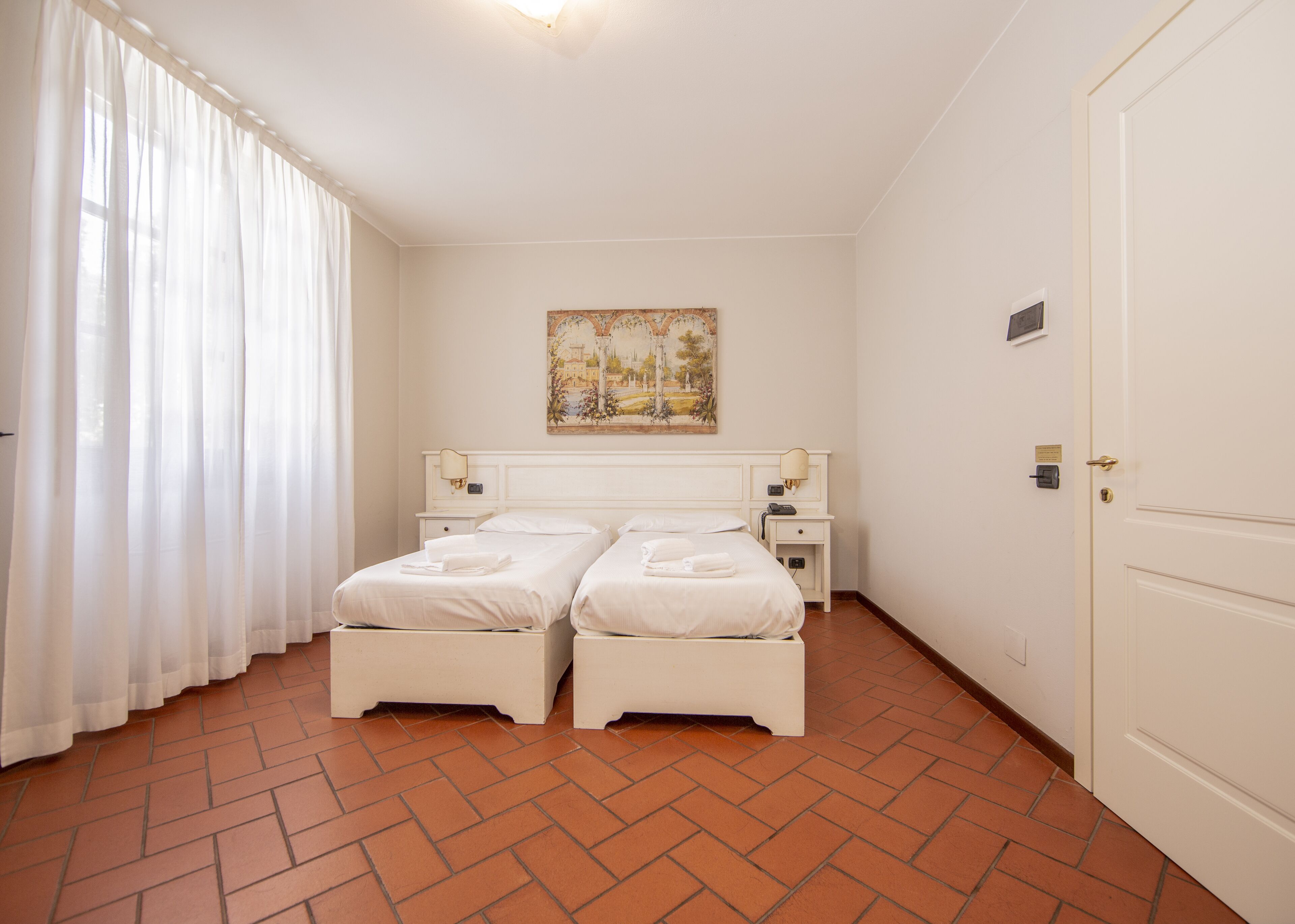 A hotel room with two single beds, a nightstand with a lamp, a painting on the wall, and a door with a lock.